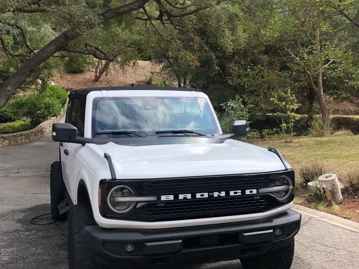 A white Ford Bronco washed and dried on a tree-lined driveway, hose still run out beside it